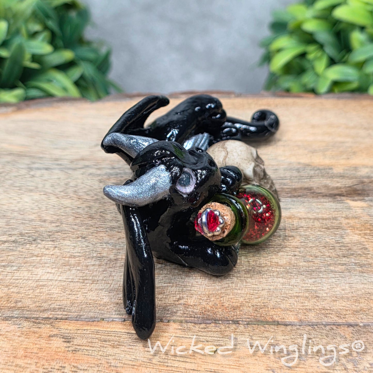 A clay dragon with a skull and potion bottle on a wood surface with greenery. Includes text Wicked Winglings