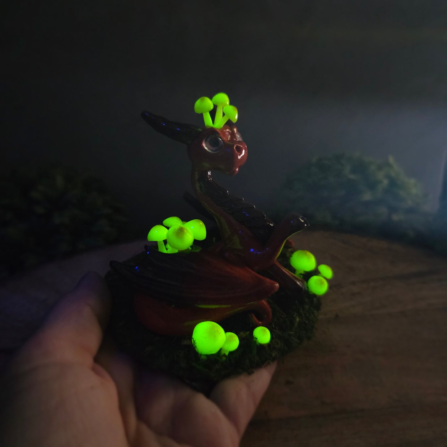 Hand holding a clay dragon figure in dark with glowing mushrooms