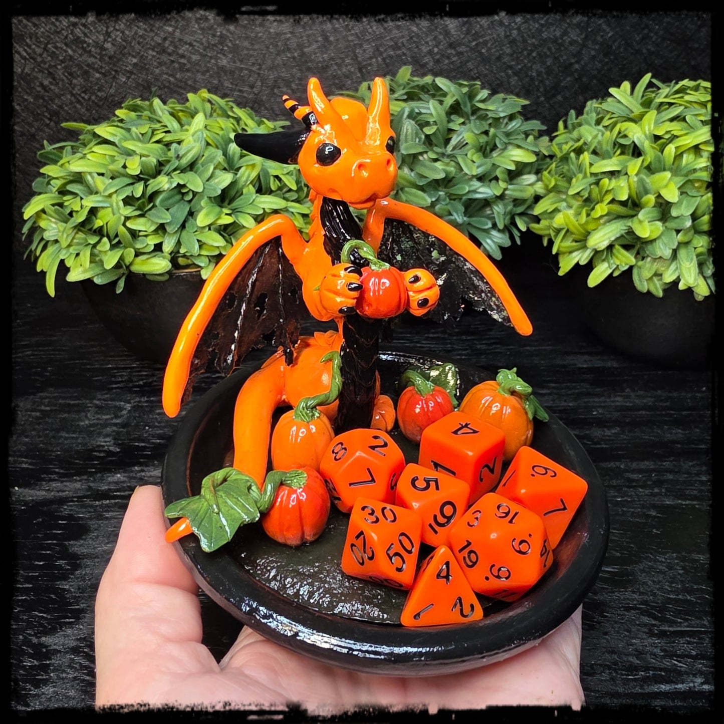 Orange dragon figurine with orange dice on a black plate against a green foliage background
