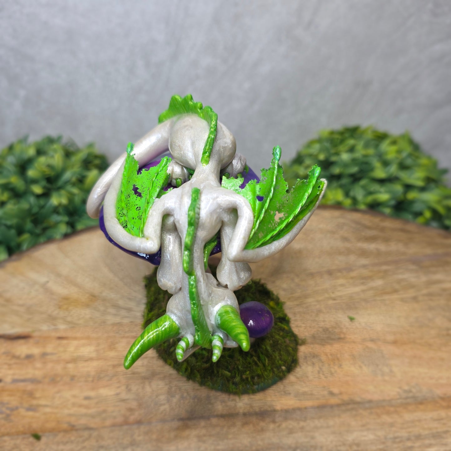 A clay dragon figurine on a mushroom on a wooden surface with greenery.