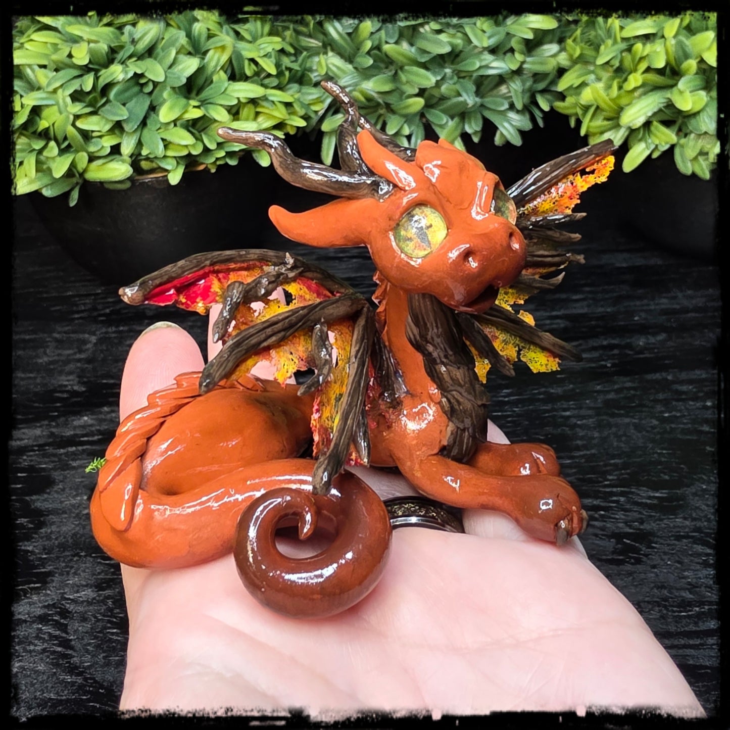 Orange dragon figurine held by a hand with green foliage in the background