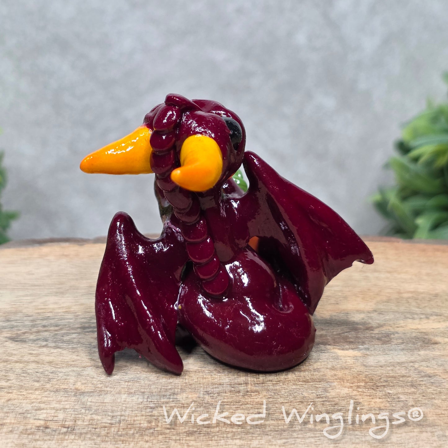 A baby clay dragon sculpture with a pumpkin against a wood background with greenery and text Wicked Winglings
