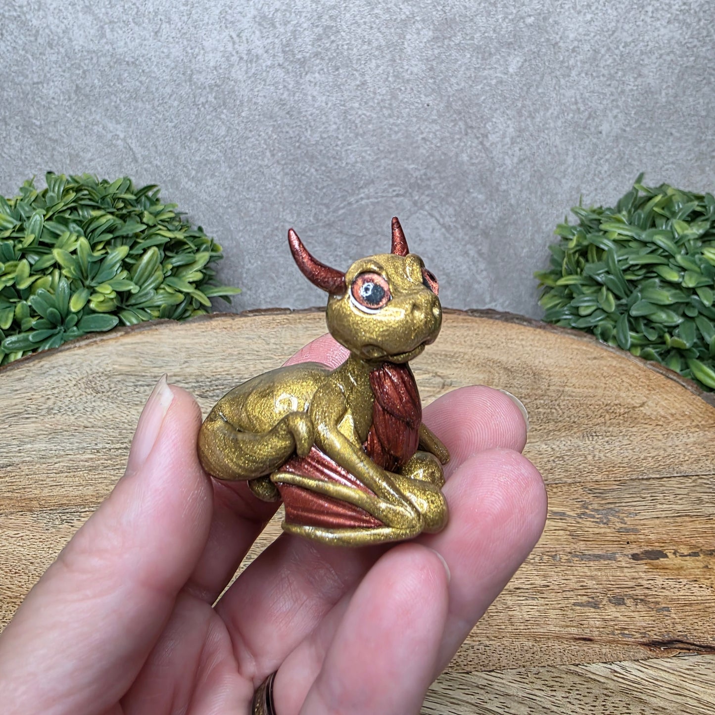 A hand holding a baby clay dragon against a wood background with greenery