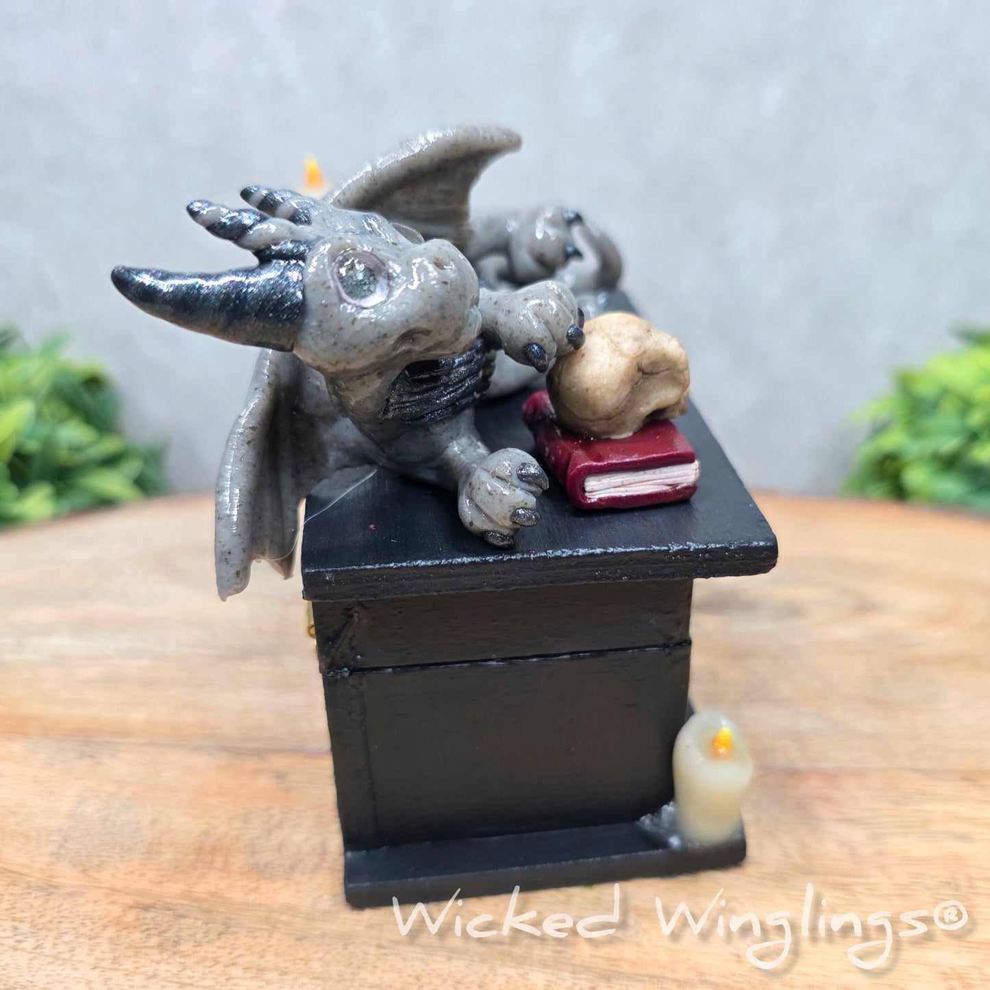 A clay dragon on a black box with candles and a skull on a wood surface with greenery. Includes text Wicked Winglings