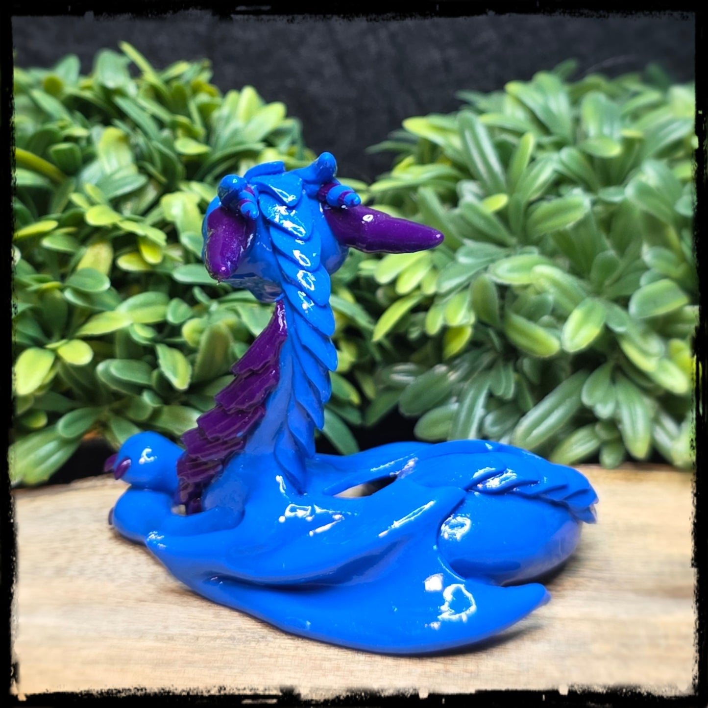 Blue and purple dragon figurine on a wooden surface with green foliage in the background