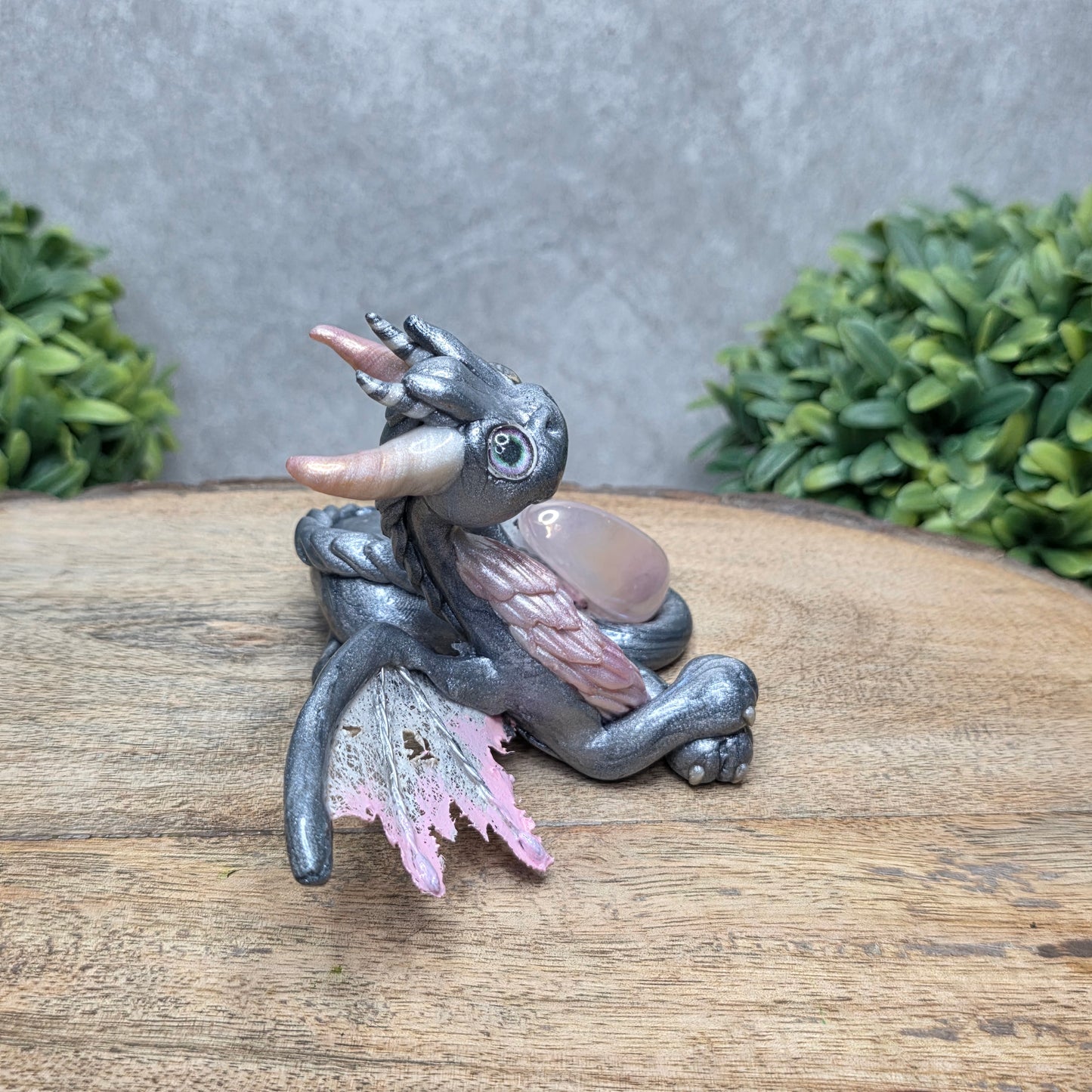 Side view of a clay dragon with a rose quartz crystal against a wood surface with greenery.