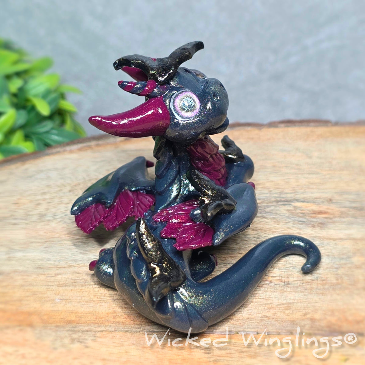 Side view of a clay dragon with bats sitting on a wood surface with greenery. Text reads Wicked Winglings.