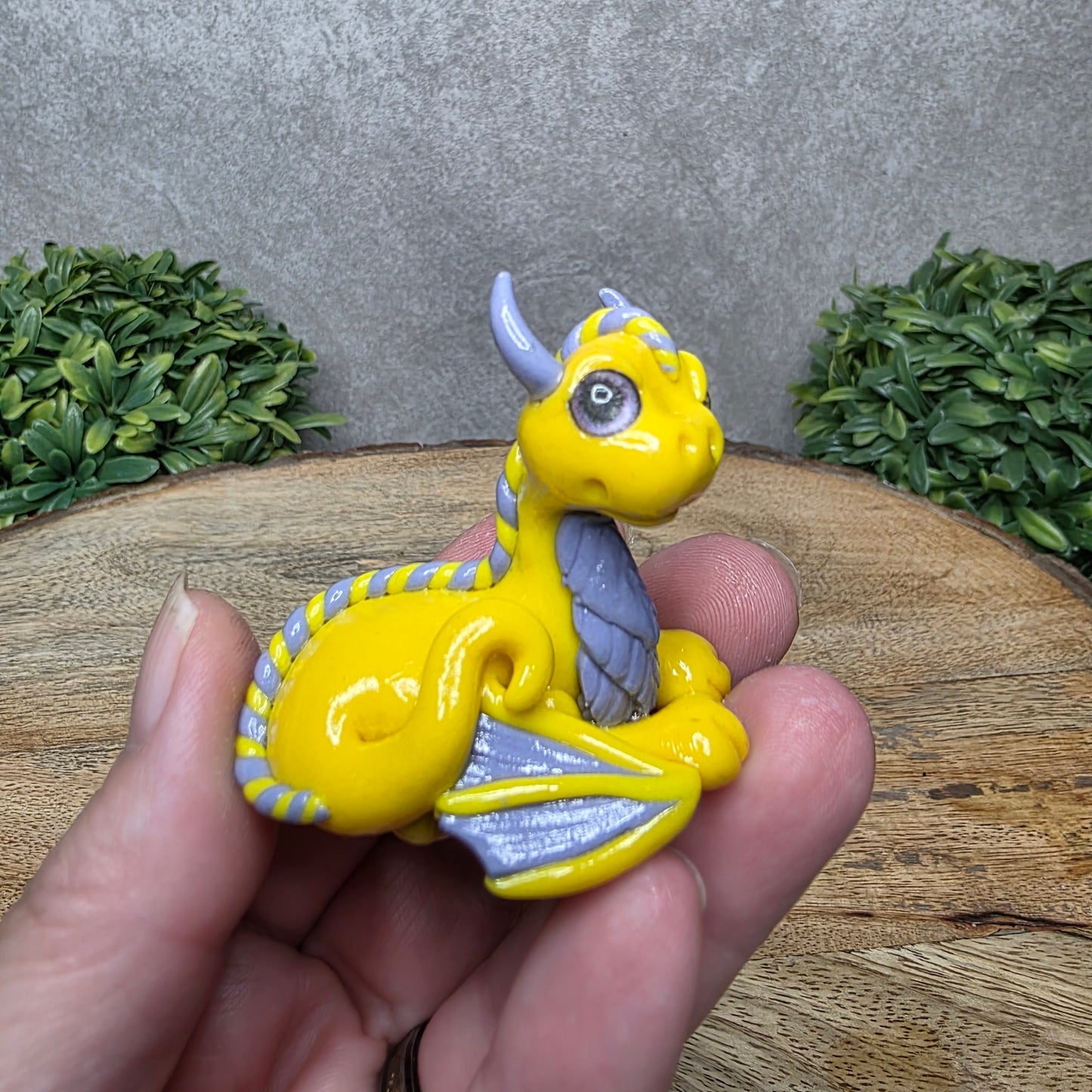 A hand holding a baby clay dragon against a wood background with greenery