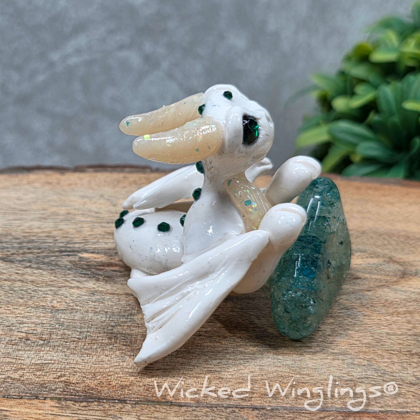 A baby clay dragon sculpture with a crackled quartz against a wood background with greenery and text Wicked Winglings