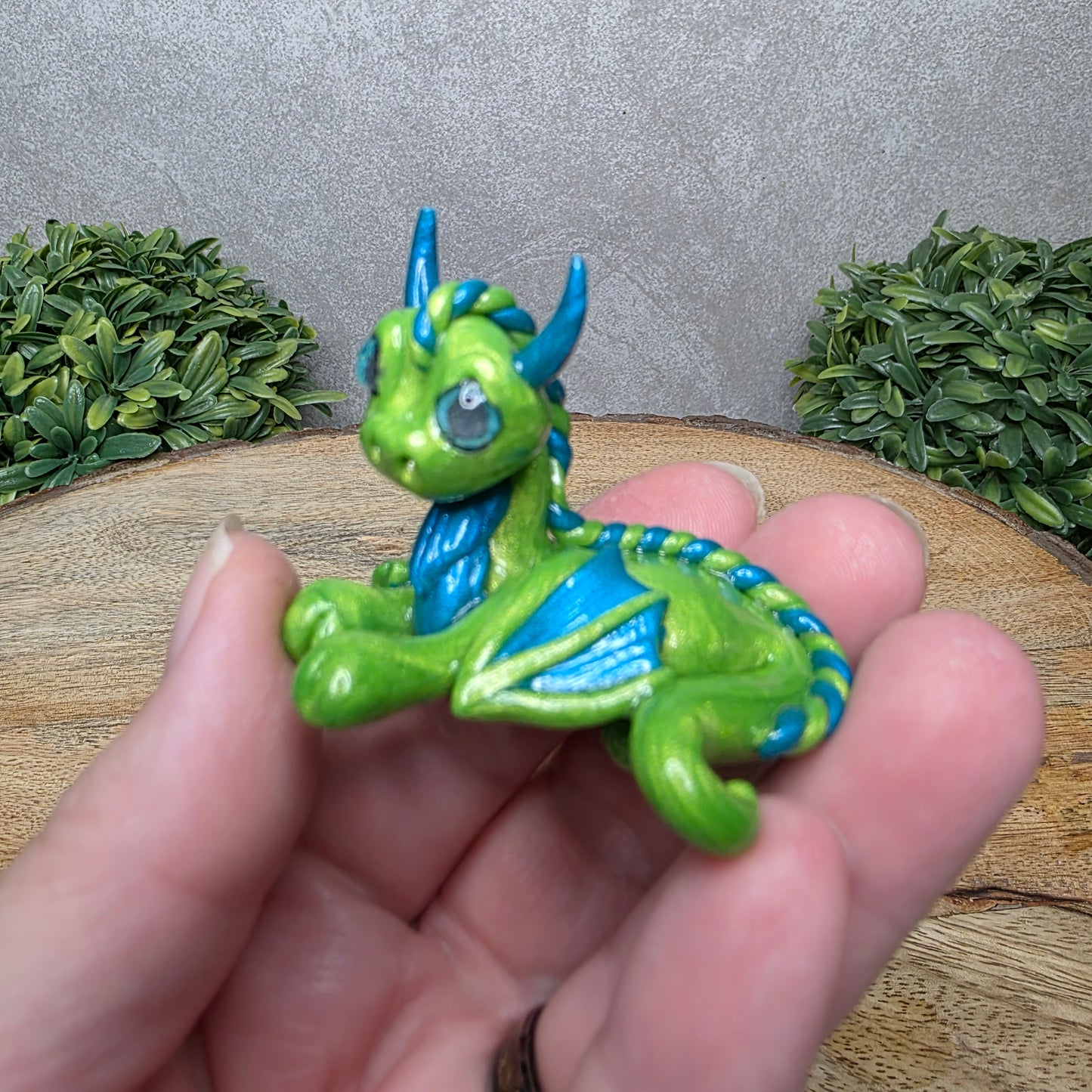 A hand holding a baby clay dragon against a wood background with greenery