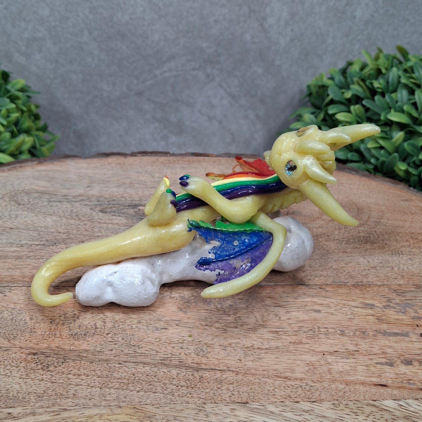 A yellow dragon with rainbow accents on a cloud sitting against a wood surface with greenery