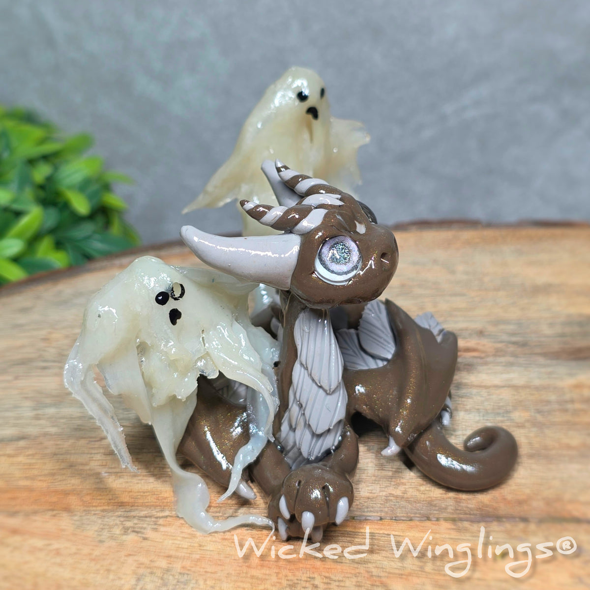 Side view of a brown clay dragon with ghosts on a wood surface with greenery. Text reads Wicked Winglings.