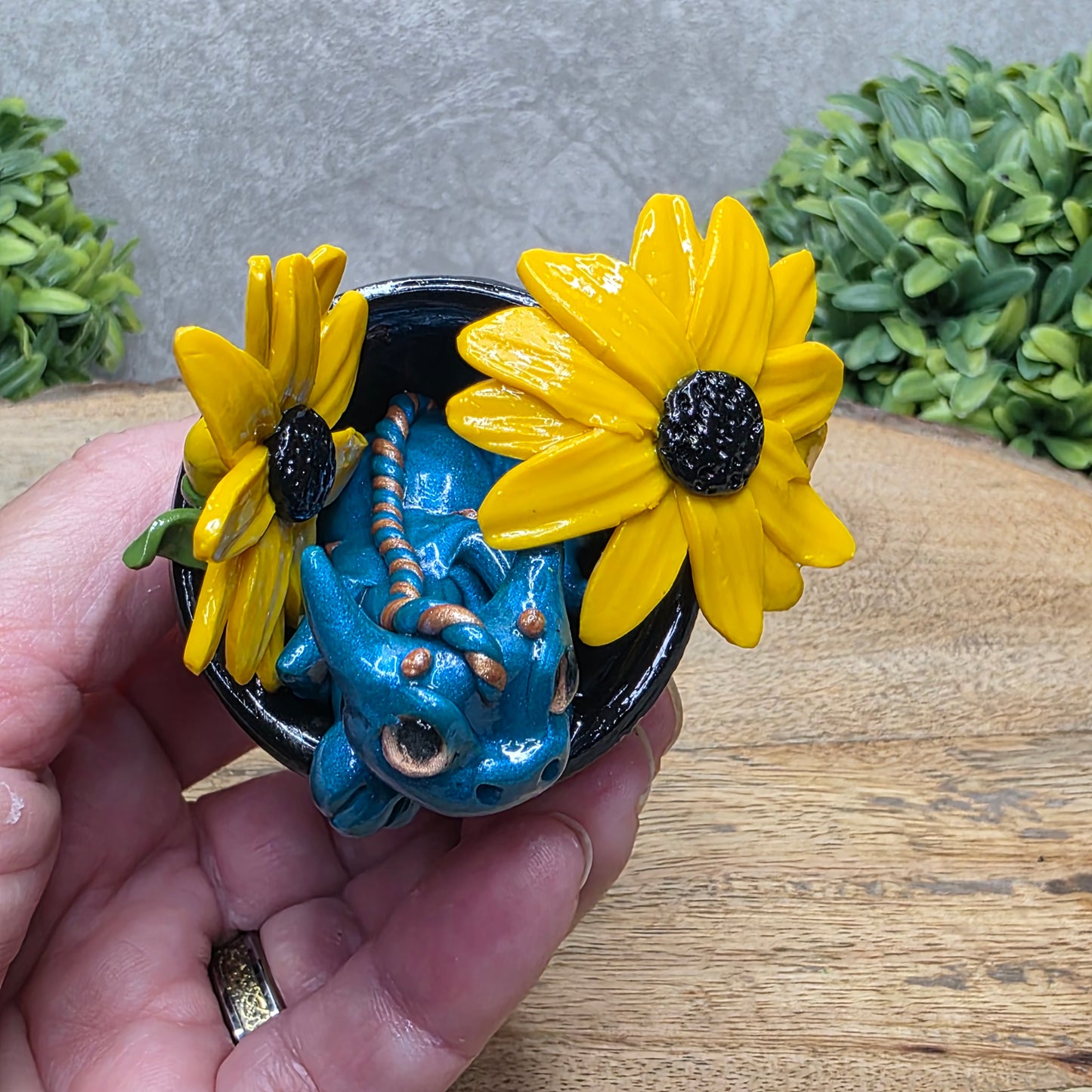Top view of a clay baby dragon in a pot with black eye susans against a wood surface with greenery.