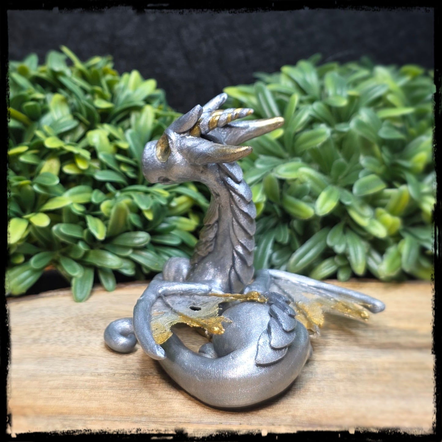Back view of a clay dragon figurine on a wooden surface with green foliage in the background
