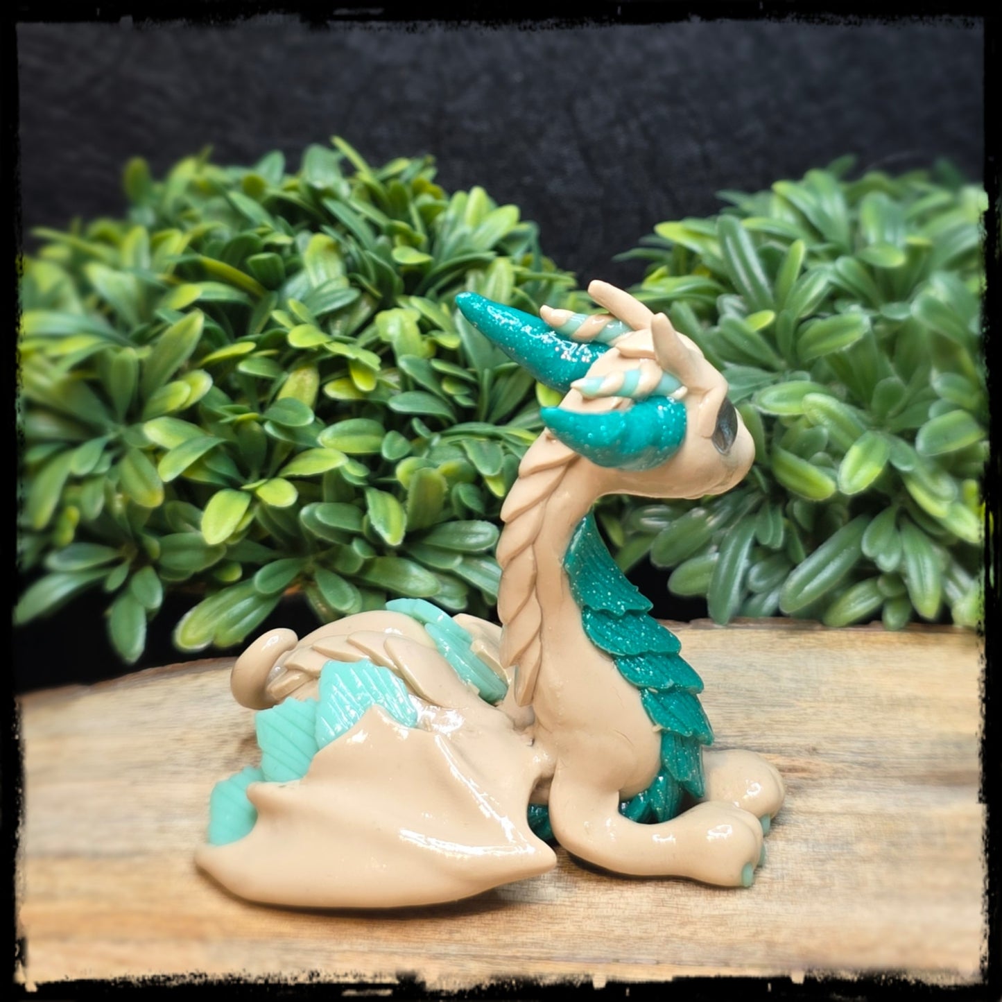 Clay dragon figurine with teal and beige colors on a wooden surface with green foliage in the background.