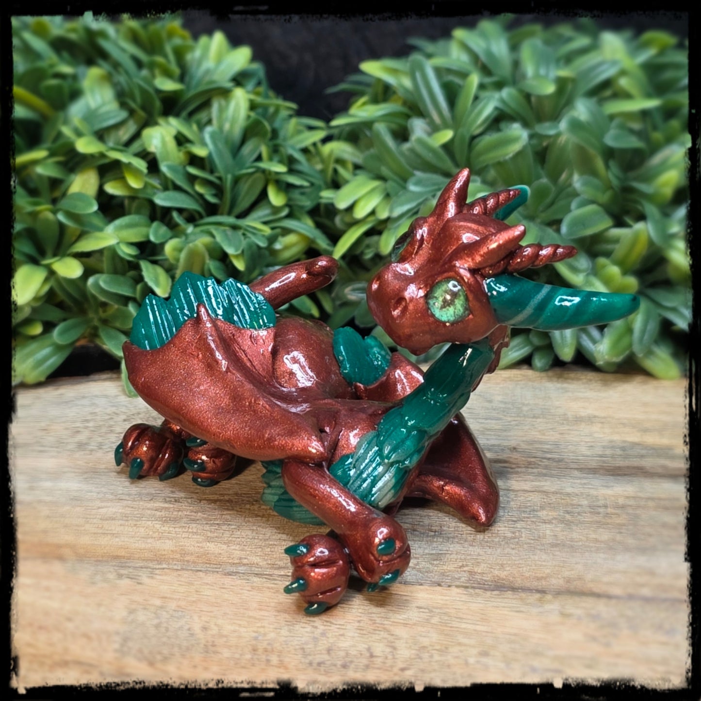 Clay bronze and green dragon figurine on a wooden surface with green foliage in the background