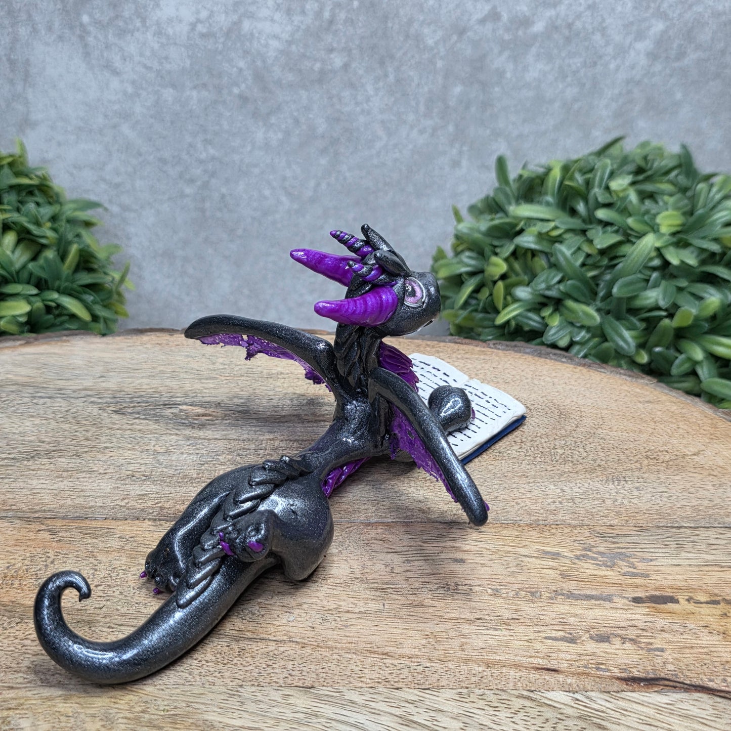 A dragon reading a book against a wood surface with greenery