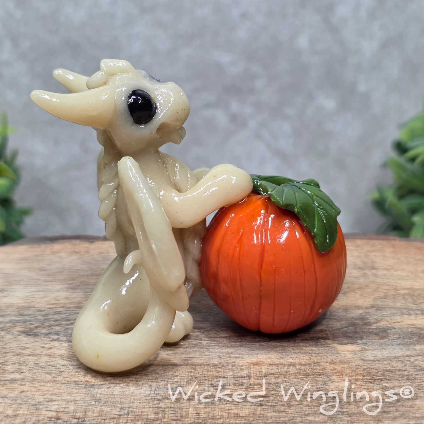 A baby clay dragon sculpture with a pumpkin against a wood background with greenery and text Wicked Winglings