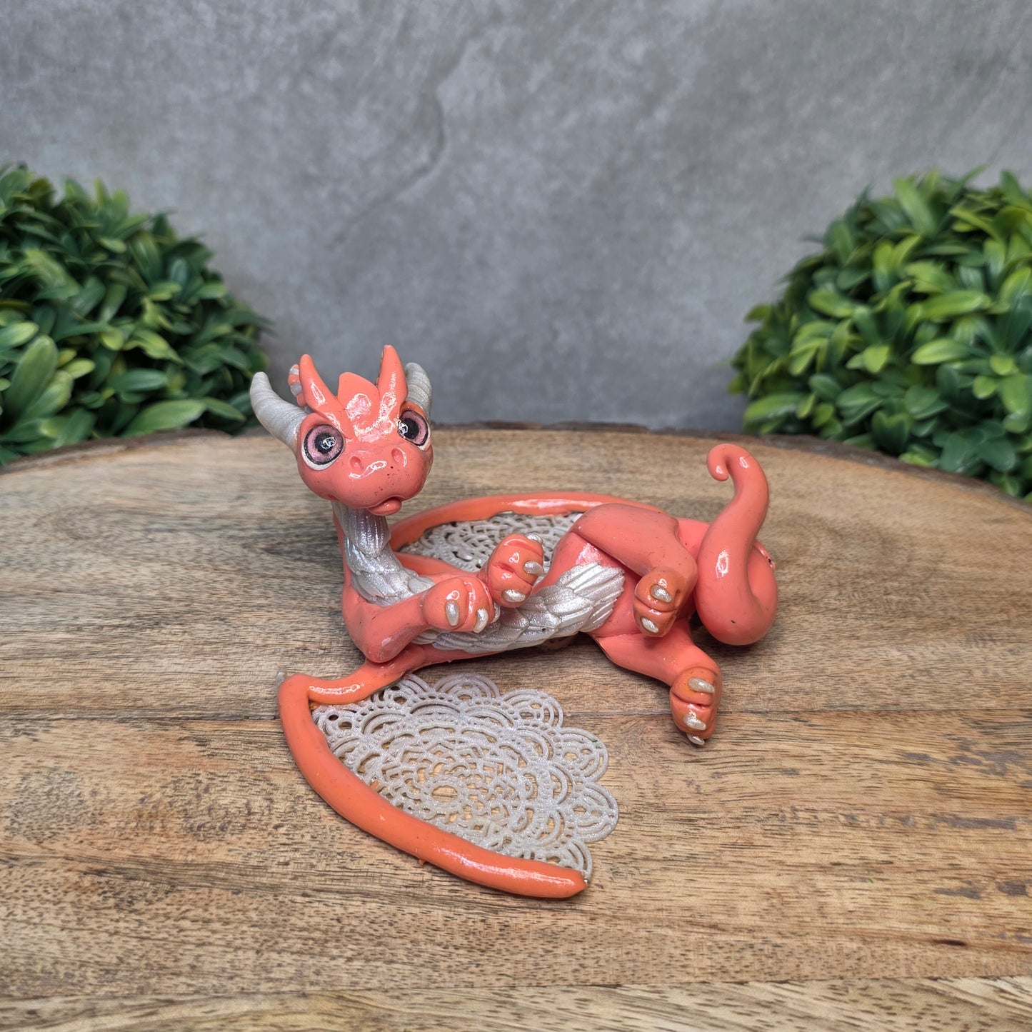 A clay dragon sculpture with lacy wings on a wood surface with greenery.