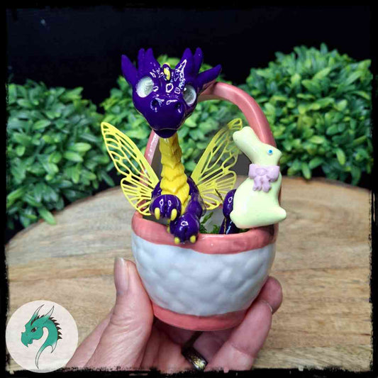 Hand sculpted multicolored dragon with yellow wings inside a ceramic basket, decorated with moss, holding a smaller white and pink bunny.