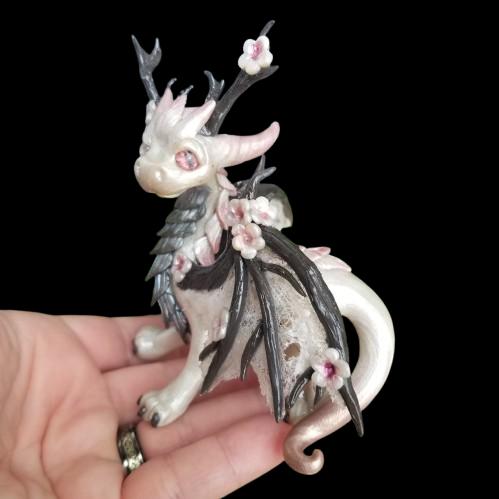 Photo features a hand holding a pearl colored clay dragon with cherry blossoms accenting horns and wings.