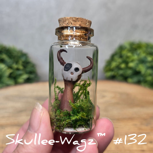 Hand holding a skull faced creature in a jar against a wood background with greenery