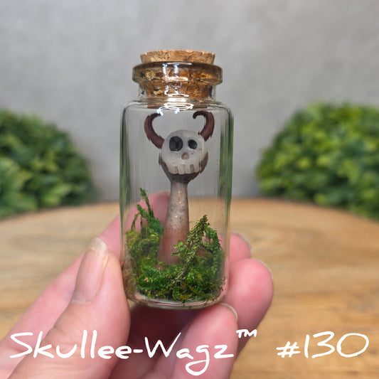 Hand holding a skull faced creature in a jar against a wood background with greenery