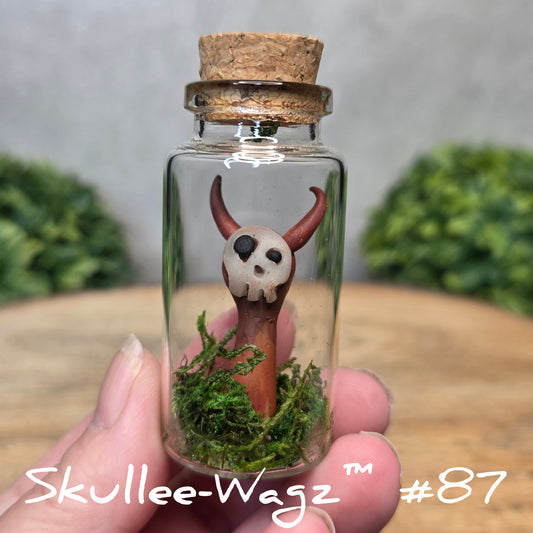 Hand holding a skull faced creature in a jar against a wood background with greenery