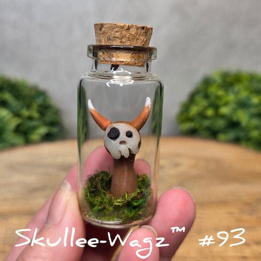 Hand holding a skull faced creature in a jar against a wood background with greenery