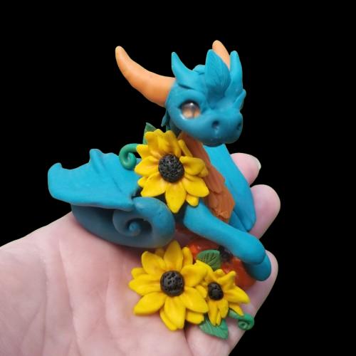 Photo features a hand holding a blue  colored clay dragon sculpture accented with sunflowers