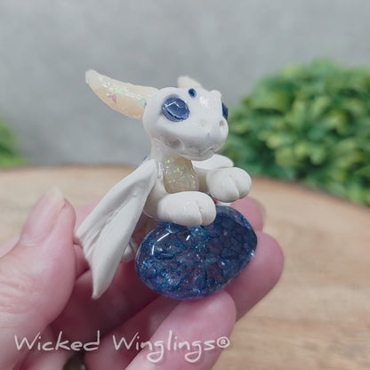 A video of a baby clay dragon sculpture with a crackled quartz against a wood background with greenery and text Wicked Winglings