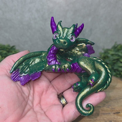 A hand holding a clay dragon against a wood background with greenery