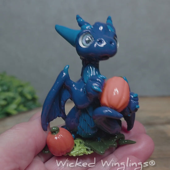 A video of a hand holding blue clay dragon with pumpkins against a wood surface with greenery. Text reads Wicked Winglings. 
