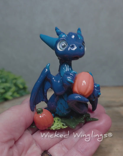 A video of a hand holding blue clay dragon with pumpkins against a wood surface with greenery. Text reads Wicked Winglings. 