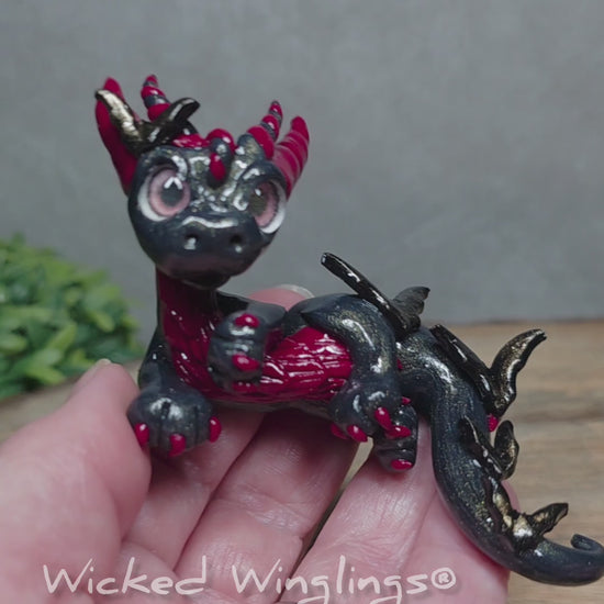 A video of a hand holding a clay dragon with bats against a wood surface with greenery. Text reads Wicked Winglings.