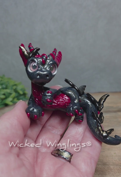 A video of a hand holding a clay dragon with bats against a wood surface with greenery. Text reads Wicked Winglings.