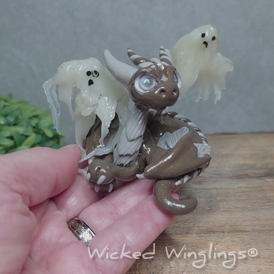 A video of a hand holding a brown clay dragon with ghosts against a wood surface with greenery. Text reads Wicked Winglings.