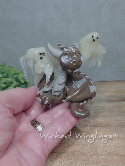A video of a hand holding a brown clay dragon with ghosts against a wood surface with greenery. Text reads Wicked Winglings.