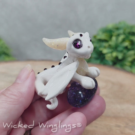 A video of a baby clay dragon sculpture with a crackled quartz against a wood background with greenery and text Wicked Winglings