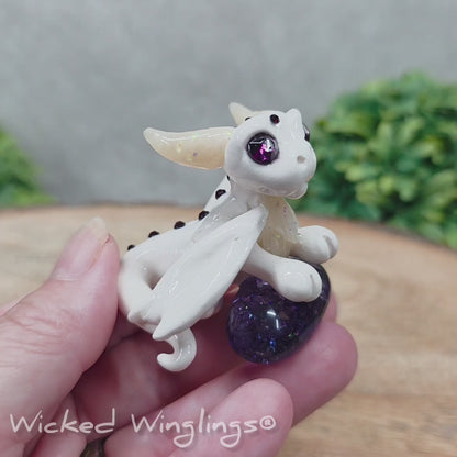 A video of a baby clay dragon sculpture with a crackled quartz against a wood background with greenery and text Wicked Winglings