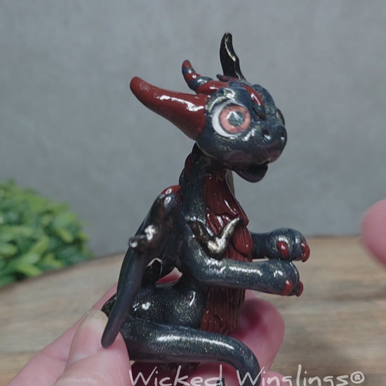 A video of a hand holding a clay dragon with bats sitting on a wood surface with greenery. Text reads Wicked Winglings.