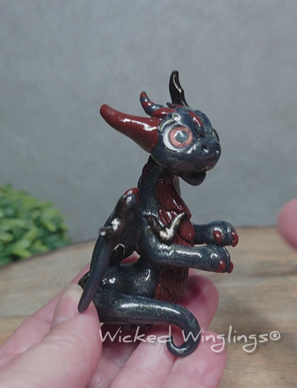 A video of a hand holding a clay dragon with bats sitting on a wood surface with greenery. Text reads Wicked Winglings.