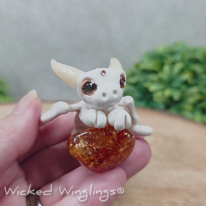 A video of a baby clay dragon sculpture with a crackled quartz against a wood background with greenery and text Wicked Winglings