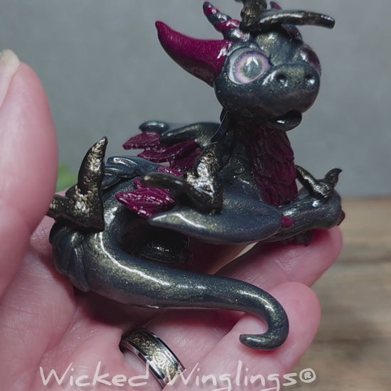 A video of a clay dragon with bats against a wood surface with greenery. Text reads Wicked Winglings.