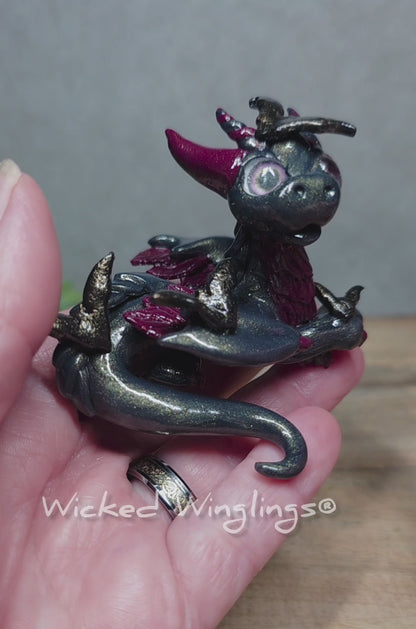 A video of a clay dragon with bats against a wood surface with greenery. Text reads Wicked Winglings.