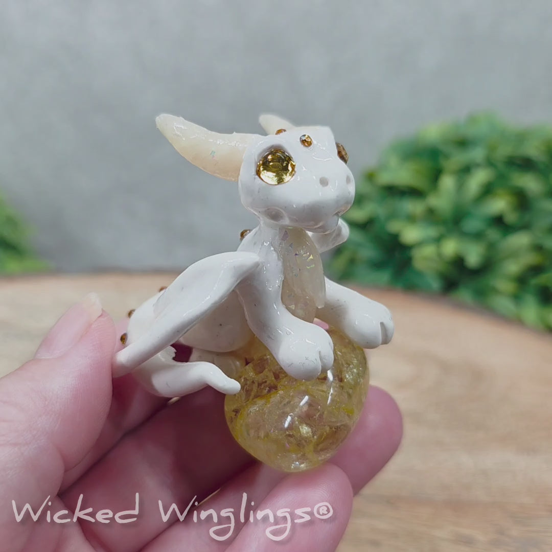 A video of a baby clay dragon sculpture with a crackled quartz against a wood background with greenery and text Wicked Winglings