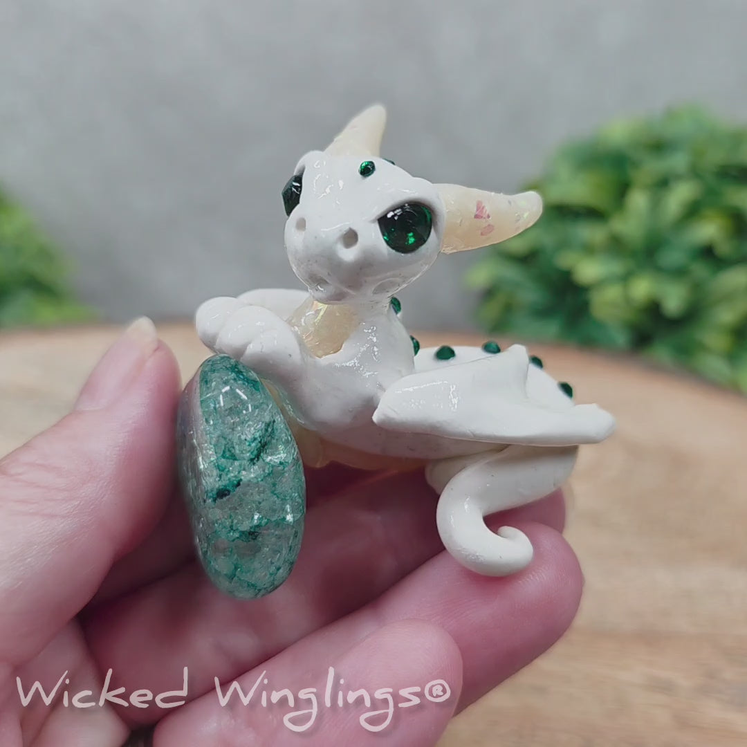 A video of a baby clay dragon sculpture with a crackled quartz against a wood background with greenery and text Wicked Winglings