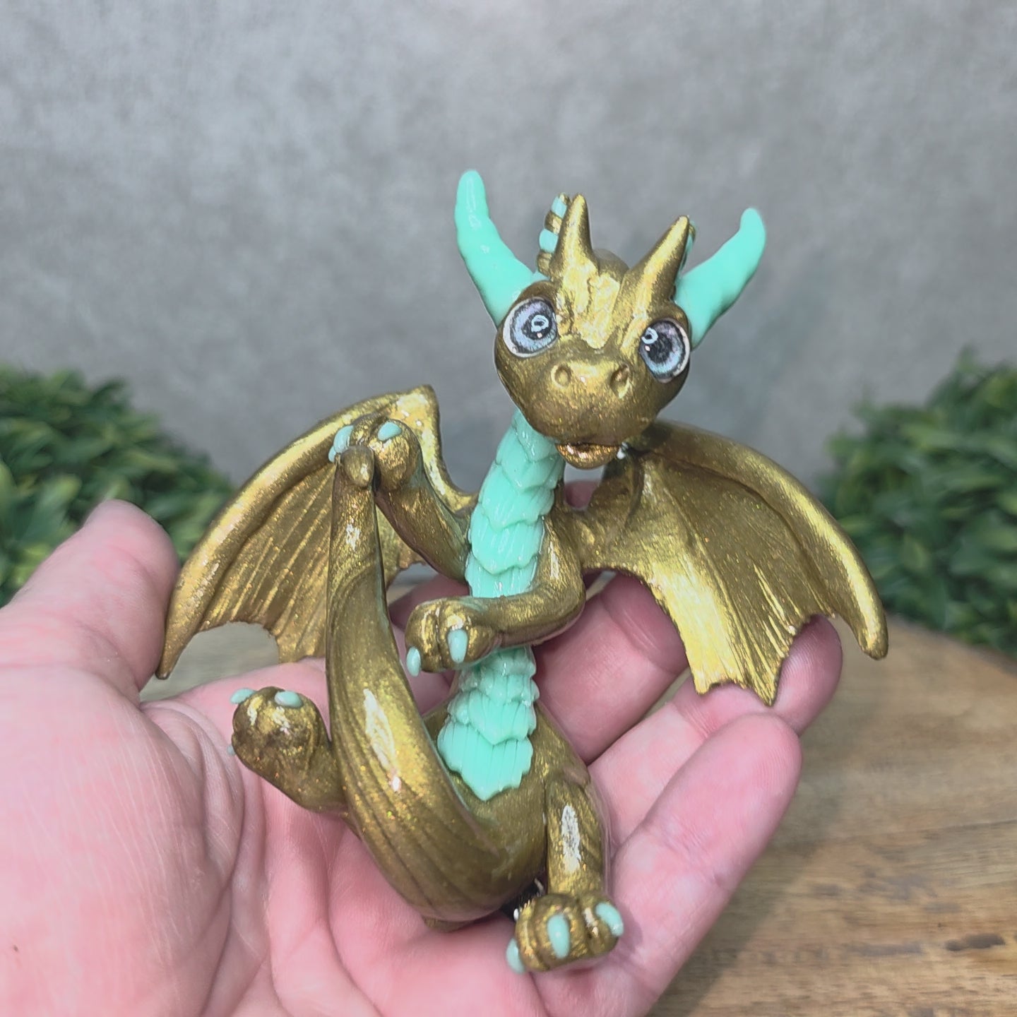 Video of A hand holding a clay dragon against a wood background with greenery