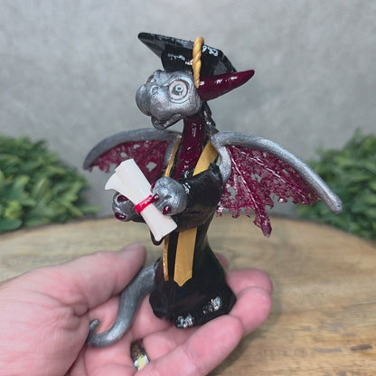 A hand holding a clay dragon in a graduation cap and gown holding a diploma
