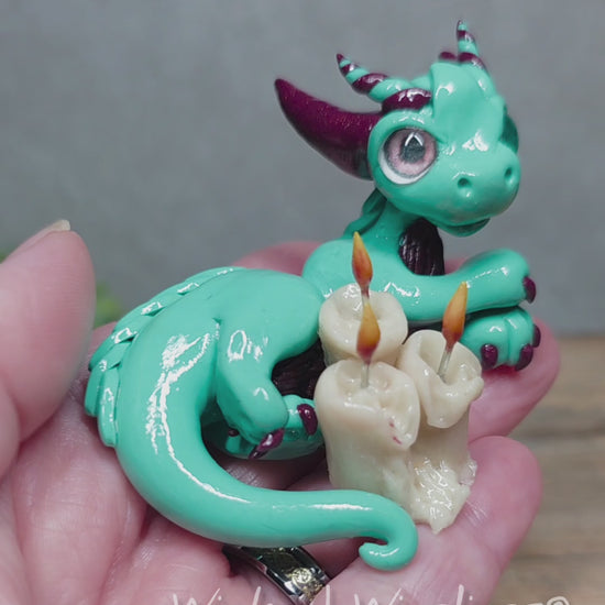 A video of a hand holding a clay dragon with candles against a wood surface with greenery. Text reads Wicked Winglings.
